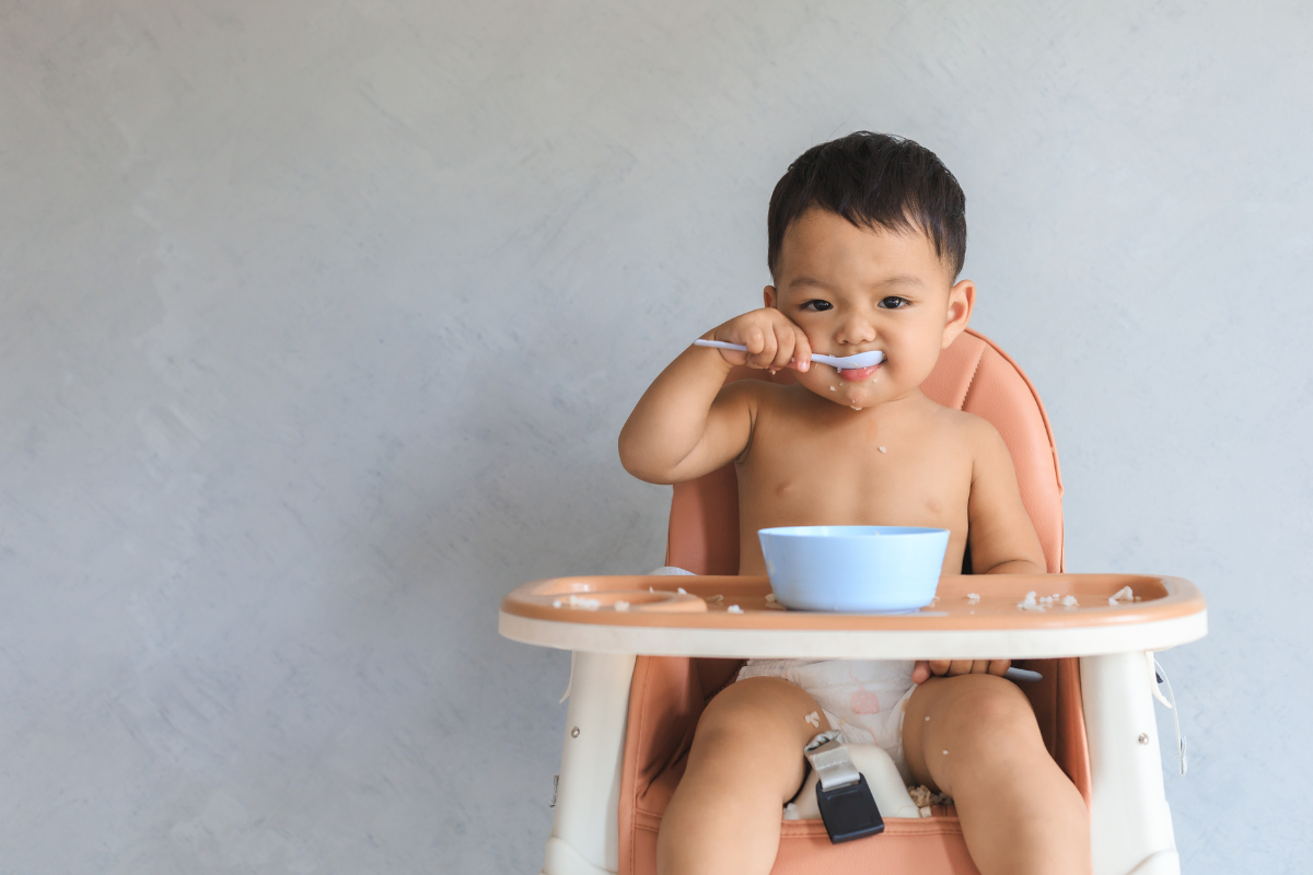 A baby boy messily eating a snack with a spoon, food smeared around his mouth and hands, enjoying the meal with playful enthusiasm.